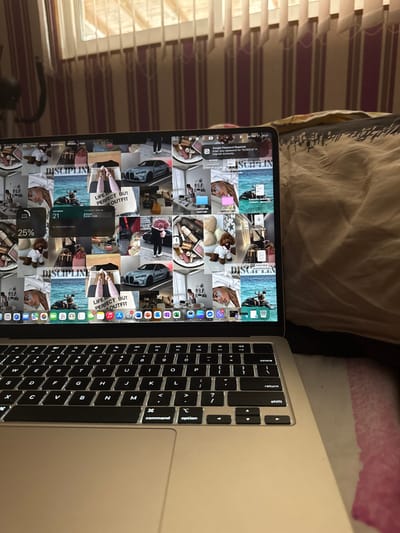 The scene depicts an open, silver-colored laptop, identified as a MacBook, positioned in an indoor setting within the city of Leshnitsa, Bulgaria. The laptop's black keyboard with illuminated white keys occupies the lower half of the frame. Its screen displays a multi-image collage wallpaper themed around luxury lifestyle, fashion, and personal care. Specific visual elements on the screen include a gray BMW M4, various designer handbags, fashion apparel (e.g., shoes, pink pants), cosmetic products, a person holding a large bouquet, a small dog in clothing, and a person on a jet ski. Overlaying the wallpaper are several system interface elements: a battery widget showing 25% charge, a calendar widget indicating 'Wednesday, Mar 27' with 'No More Events Today,' a Google Chrome notification prompting for a password related to 'BG230213' to access saved passwords, and the menu bar displaying 'Wed Mar 27 16:31'. A dock with various application icons is visible at the bottom of the screen.
The laptop rests on a surface partially covered by a light-colored blanket or sheet with subtle pink and white stripes. To the immediate right of the laptop, a rumpled beige or light brown blanket/duvet is visible, suggesting the laptop is positioned on a bed. The background features a window covered by vertical blinds or striped curtains, allowing ambient light to illuminate the upper portion of the room. The wall behind the laptop exhibits a vertical striped pattern in shades of maroon or pink and white/off-white. A dark, curved object, possibly part of exercise equipment, is partially visible and out of focus in the upper left background. No individuals are present or actively interacting with the laptop within the visible frame.