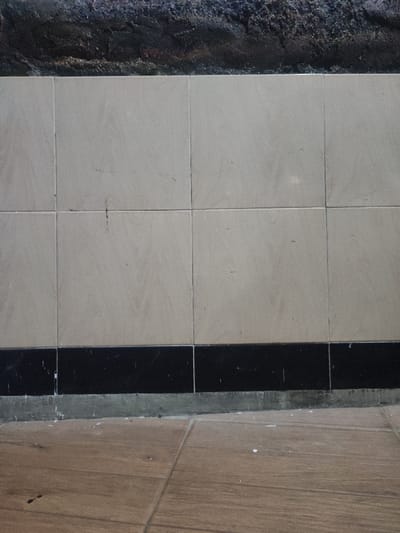 The image shows a wall made of white tiles with a black tile trim at the base. The wall is made of 6 large, white tiles and one row of black tiles. The tiles are slightly worn and scuffed, indicating use. The wall is against a brown tile floor. The image was taken in a building located in Kecamatan Prigen, Indonesia.