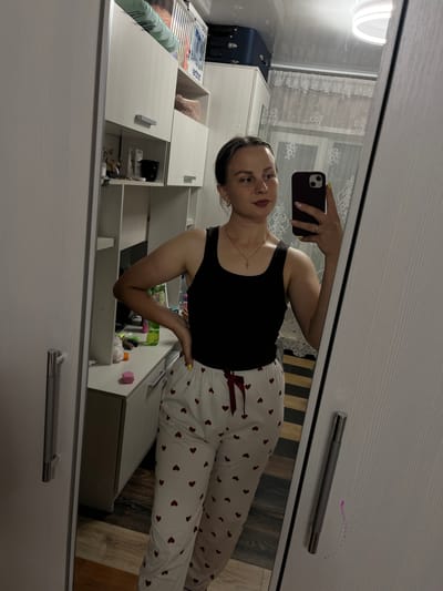The image shows a woman standing in what appears to be a bedroom or dressing area. She is taking a mirror selfie, holding a smartphone in her right hand. The woman is wearing a black tank top and white pajama pants with a red heart pattern. She has on a gold necklace with a cross pendant and hoop earrings. Her hair is pulled back, and she is wearing lipstick.
In the background, there is a large wardrobe with mirrored doors, reflecting the room. Part of the room visible in the mirror includes shelves with various items, including what looks like storage boxes, bottles, and decorative objects. There is also a glimpse of a window with a patterned curtain.
The scene is set within an indoor environment, likely a home. The presence of a wardrobe and bedroom-like items suggests a personal space. While the exact location within Balezino, Russia cannot be definitively determined from the visual information alone, the overall setting is consistent with a residential interior in any urban or suburban area.