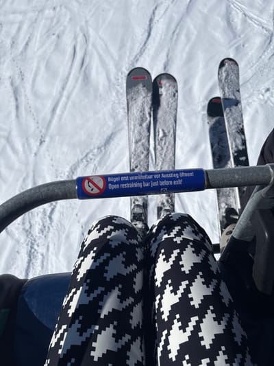 The image captures a downward-angled, first-person perspective from a ski chairlift in Zürs, Austria, under bright, clear daylight conditions. The subject's lower legs are prominently featured, clad in black pants with a white pixelated houndstooth-like pattern, resting on a blue upholstered chairlift seat. A grey metallic restraining bar spans horizontally across the frame in the mid-ground. Attached to this bar is a blue rectangular sign displaying white text in German ("Bügel erst unmittelbar vor Ausstieg öffnen!") and English ("Open restraining bar just before exit!"), accompanied by a red circular 'no standing' pictogram and a small 'Doppelmayr' logo. Below the restraining bar, four skis are visible, partially covered in snow. Two skis belong to the primary subject, positioned directly ahead, and two additional skis are located to the right, suggesting a second occupant on the chairlift. The vast snow-covered ground beneath the chairlift exhibits numerous ski tracks and faint animal footprints.