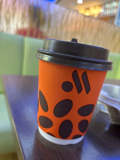 An orange coffee cup with a black lid and black polka dots rests on a wooden table. The cup features a logo that appears to be two stylized lines with a dot. In the background, out of focus, are green and light-colored surfaces, possibly seating or walls in a cafe. The overall scene suggests an indoor setting, likely a cafe or eatery. There is no identifiable information about the city of Shendzhii, Russia, in the image.