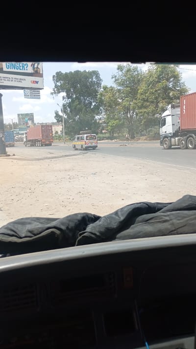 The image shows a road scene viewed from inside a vehicle. A billboard advertising "GINGER?" with health warnings is visible. The landscape features trees, a white and yellow minibus, and large trucks carrying shipping containers. The dashboard of the vehicle occupies the bottom portion of the image, with a dark cloth draped over it. The view suggests the vehicle is either stopped or moving slowly at an intersection.
