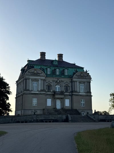 The image shows a Baroque palace, likely in Kongens Lyngby, Denmark, during a clear, possibly late afternoon or early evening, given the soft light. The building is grand and ornate, featuring a distinctive green copper roof with dormer windows and prominent chimneys. The facade is constructed of stone with decorative elements, including sculptures and relief work. Multiple stories are visible, characterized by numerous windows with white frames.
The palace is situated on a slight incline, with a wide stone staircase leading up to the entrance. To the right of the building, a gravel path runs alongside a well-maintained grassy area. The setting suggests a park or formal garden surrounding the palace. The overall atmosphere is one of grandeur and historical significance, with the clear sky and soft lighting contributing to a serene and perhaps slightly nostalgic mood. No people are visible, and the scene is peaceful. No text is present in the image.