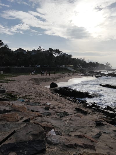 The image captures a view of a beach in Phú Thủy, Vietnam. In the foreground, there are scattered rocks and sand, with some visible pieces of trash like plastic bags. The midground shows a sandy beach with a few people walking and interacting. Further back, the ocean meets the shore with gentle waves. Along the coastline, there is a line of trees and houses, suggesting a residential or resort area. The sky is partly cloudy with bright sunlight filtering through, indicating it's likely daytime, possibly late afternoon or early morning. A road with some parked vehicles is visible behind the beach area.