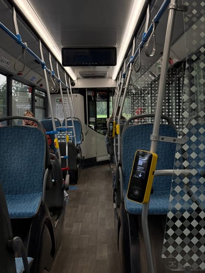 The image shows the interior of a bus in Yakovlevskoye, Russia, looking down the aisle towards the front. The seats are blue with a checkered pattern, and there are metal poles and hand straps for passengers. A woman with blonde hair is visible on the left, talking on her phone. On the right side of the aisle, a yellow card reader is mounted on a pole. The floor is dark wood-grain.