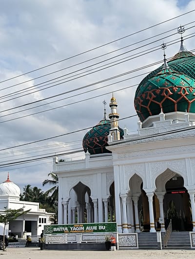 The image captures a ground-level view of Masjid Baitul Yaqin Alue Bu, a mosque located in the city of Aluebutuha, Peureulak Barat district, Indonesia. The mosque's architecture is predominantly white, featuring detailed arched entryways supported by columns. Two green and gold domed structures are visible atop the building, adorned with intricate patterns and topped with crescent moon symbols. Power lines stretch across the sky above the mosque. The entryway has steps and a stainless-steel handrail that leads into the interior. To the left of the main building, a smaller white structure with a red-topped dome stands. A sign in front of the mosque displays its name and location. A car is parked near the left side of the image. The sky is overcast with cloudy conditions.