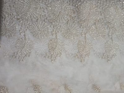 The image shows a delicate white or off-white fabric, possibly tulle or netting, adorned with intricate embroidery. The embroidery consists of small, shimmering floral or circular patterns, arranged in a loosely cascading design along the top portion of the fabric. The fabric appears textured, with a subtle sparkle due to the metallic thread or beads used in the embroidery. The background is a blurred, undefined white, creating a soft, ethereal feel.