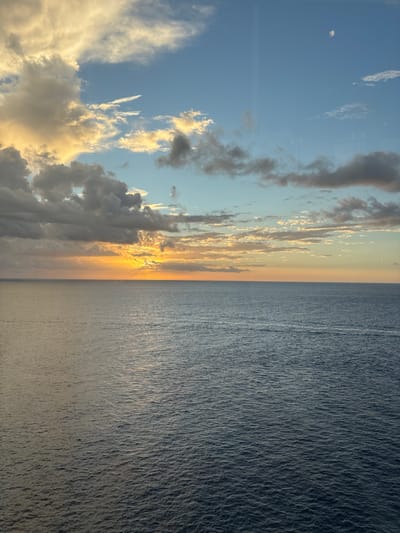 The image displays an expansive seascape and sky observed from the vicinity of San Miguel de Cozumel, Mexico, during either sunrise or sunset. The horizon line, extending horizontally across the frame, separates the water from the sky. The lower two-thirds of the image consist of the ocean's surface, characterized by dark blue and grey tones and fine ripples. A path of warm, orange-yellow light reflects on the water, emanating from the sun's position near the horizon.
The upper third of the image is occupied by the sky, transitioning from a light blue at the top to vibrant orange and yellow hues where the sun is partially visible just above the horizon, slightly left of center. The sky contains multiple cloud formations: dense, layered clouds with golden illumination are present on the left, while sparser, smaller clouds are scattered throughout the mid and upper right sections. A distinct crescent moon is visible in the upper right corner of the sky. Faint, vertical linear reflections are discernable across the sky, suggesting the image was captured through a window. No individuals, vessels, or man-made structures are present within the visible scene.
