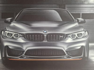 This is a medium shot, eye-level photograph of the front of a silver BMW M4. The car is positioned centrally in the frame, facing directly towards the viewer. The background appears to be a garage or industrial setting, with a corrugated metal door behind the car, rendered in shades of gray.
The car's design is aggressive and sporty. Prominent features include the distinctive kidney grille with vertical slats, flanked by angular headlights with circular LED daytime running lights. The front bumper has large air intakes with honeycomb grilles, and a thin, bright orange accent line runs horizontally across the lower part of the bumper, adding a vibrant contrast to the metallic silver paint. A small "M" badge is visible on the right side of the kidney grille.
The lighting is even, suggesting an indoor setting or overcast conditions, with no harsh shadows. The overall mood is one of power and performance, highlighting the vehicle's aesthetic appeal. There is no visible text other than the "M" badge on the grille. The image appears to be a professional studio shot or a well-composed photograph taken in a controlled environment.