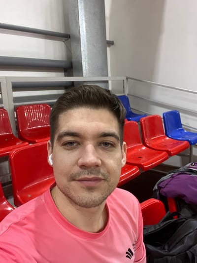 The image is a selfie-style portrait of a man taken indoors, likely within a sports facility or arena. The man, who appears to be in his twenties or thirties, is positioned in the foreground, looking directly at the camera with a neutral to slightly smiling expression. He has short, dark hair, light-colored eyes (blue or grey), and a short, well-groomed stubble beard. He is wearing a bright, solid pink short-sleeved athletic shirt, which has a black Adidas logo featuring the three stripes and text visible on the upper right shoulder/collar area. A white earbud is clearly visible in his left ear.
The background primarily consists of rows of empty stadium-style seats. Most of the visible seats are a vibrant red, with a few blue seats interspersed, suggesting a pattern or specific section. Behind and above the seats, the wall is white, and industrial elements are apparent, including grey metal beams, support structures, and several grey pipes or ducts running along the upper part of the wall and ceiling. In the upper right corner of the image, a dark-colored bag or jacket is resting on one of the red seats. The lighting is bright and even, indicative of indoor artificial illumination. The overall scene suggests a moment of rest or waiting in a sports venue.
