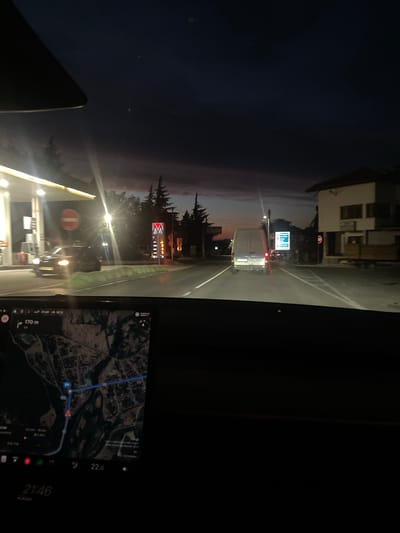 The image is taken from inside a vehicle at dusk, looking forward. The vehicle is on a road with streetlights and signs. To the left, there is a gas station with bright lights. A black car is visible at the gas station. Further down the road, a white van is driving away from the camera. On the right side of the road, there is a building and a road sign. The dashboard of the vehicle shows a map navigation system, the time 21:46, and a temperature of 22.0 degrees Celsius. The sky is dark with hints of purple and orange, indicating sunset or twilight. The overall scene depicts a journey through a town or city at the end of the day. The location is identified as Vetren Dol, Bulgaria.