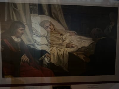 The image shows a painting of a woman lying in bed, appearing ill. She is wearing a light-colored gown and a head covering, with a medal around her neck. Two figures are attending to her. On the left, a person in dark clothing with red fabric draped over their shoulder looks down at the woman. On the right, another person, partially obscured by darkness, leans over a table next to the bed. The setting appears to be a dimly lit room with a canopy over the bed. Below the painting, text identifies the artwork as "Isabel the Catholic dictating her Will" by Eduardo Rosales. The text also indicates "SPANISH PAINTING" and mentions the city of Mazarrón, Spain, suggesting the painting may be displayed there.