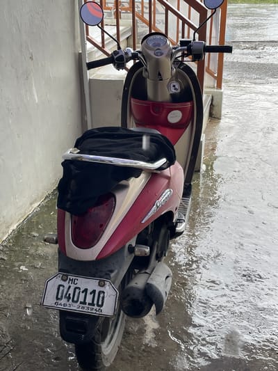 An outdoor, medium-shot, eye-level, full-color, high-angle photographic image taken from the rear of a parked motorcycle. The motorcycle is a Yamaha Mio, a scooter-style motorcycle that is red and cream colored. It is parked on a wet, concrete surface. There is a black mesh bag draped over the rear seat of the motorcycle. The motorcycle's license plate is visible and reads "MC 040110". The background shows a wall on the left, and a set of stairs with a railing on the right, suggesting an outdoor area attached to a building. The overall environment appears to be outdoors on a cloudy day, with visible puddles and wet surfaces indicating recent or ongoing rain. The location is confirmed to be San Jose del Monte, Philippines, due to the presence of a "MC" (Motorcycle) designation on the license plate, which is typical for Philippine motorcycle plates.