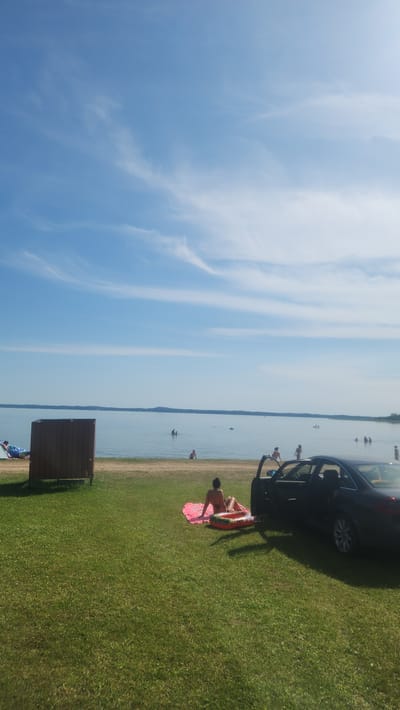 The image depicts a sunny day at a lakeside in Tilīši, Latvia. The scene is divided into a bright blue sky with wispy clouds and a serene body of water stretching towards a distant, hazy horizon. A grassy area borders the water, with a dark-colored car parked on the grass.
Several people are visible. In the foreground, a person is lying on a pink inflatable raft on the grass, facing away from the camera. Near the water's edge, a few other individuals can be seen, some partially submerged in the water, suggesting they are swimming or wading. Further out in the lake, more people are present, enjoying the water.
A small, brown wooden structure, possibly a changing room or a storage unit, is situated on the grassy area closer to the water. The overall atmosphere appears relaxed and leisurely, indicative of a pleasant summer day spent outdoors by the water. The bright sunlight suggests it is midday or early afternoon. No visible text is present in the image.