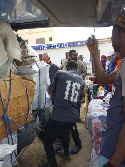 The scene depicts an outdoor commercial area in Unguwa Uku Kauyen Alu, Nigeria. In the foreground, the rear hatch of a dark-colored vehicle, likely a van or SUV, is open, revealing a tightly packed cargo area. The cargo consists of multiple large sacks (some white woven, others patterned), cardboard boxes, and bundles secured with blue ropes. A man wearing a dark grey t-shirt with the number "16" printed in white on the back, and dark trousers, is positioned centrally, bending over towards the cargo within the vehicle. He wears light-colored slide sandals. To his left, a bald man in a dark jacket looks downwards. To his right, a man in a light-colored long-sleeved shirt smiles towards the right. Partially visible in the extreme foreground right, a man in a blue sleeveless top and a patterned cap holds onto a strap from the vehicle's interior. On the ground to the right of the vehicle, additional bundled items are stacked. In the background, a light-colored building with a blue sign reading "BIS KITCHEN Eat & Take Away" is visible. Several people are gathered in the background, including two women wearing hijabs, one holding an infant, and other men. The ground surface is unpaved and appears dusty. The overall lighting suggests clear daylight.