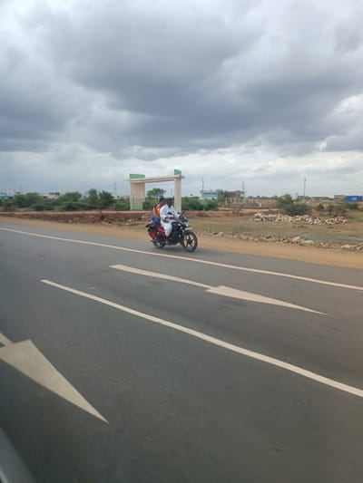 This media file captures a scene on a road in Orvakal, India. The primary subjects are two individuals riding a motorcycle. The person in the front is driving, and the person behind is a passenger. They are both wearing light-colored clothing. The motorcycle appears to be red, with a red bag or cargo strapped to the back.
The scene is set on a paved road with white lane markings. The road appears to be a two-lane highway. To the right of the road, there is a dry, somewhat barren landscape with scattered rocks and some vegetation. In the background, there is a noticeable architectural structure that resembles an archway or gate, painted in light green and white. Beyond that, there are some buildings and trees, suggesting a semi-urban or rural environment.
The weather appears overcast, with a heavy, gray cloud cover dominating the sky. This suggests it might be a cloudy day, possibly before or after rain. The lighting is diffused, and there are no strong shadows, consistent with an overcast sky. The time of day is not clearly indicated, but the muted light suggests it is not the brightest part of the day. The overall mood of the image is functional, capturing a moment of travel along a road in a rural Indian setting. There is no visible text in the image.