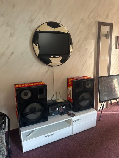 The image depicts a living room in Brca, Montenegro, with a distinct football-themed television above a white entertainment stand. On the stand are two large black Sony speakers, each adorned with a red and yellow scarf. A black DVD player and remote are also visible. To the right, an easel holds a framed chalkboard with a drawing. A full-length mirror is mounted on the wall behind the easel. The wall has a textured finish in muted tones, and the floor is covered in a dark carpet.