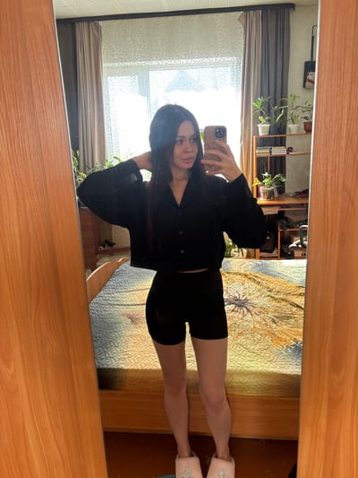 The image is a mirror selfie of a woman with long dark hair. She is wearing a black button-down shirt and black shorts, and pink slippers. She is holding a cell phone in her hand. Behind her, the reflection in the mirror shows a window with sheer curtains, a shelf with plants, and a bed with a patterned comforter. The mirror is framed with a wooden border. The location is an indoor residential setting, but there is no indication of the city of Dubrova, Russia in the image.