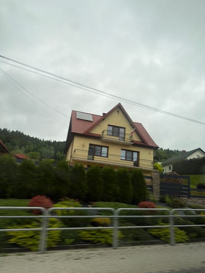 The image captures a cloudy day scene in Wilkowisko, Poland, featuring a two-story yellow house with a red tiled roof. The house has multiple windows and balconies, and solar panels are visible on the roof. The property is surrounded by lush green trees and bushes, with a prominent row of dark green evergreen trees in the foreground. A silver metal railing runs horizontally across the lower part of the frame, partially obscuring the view of the garden and a paved walkway. In the background, more trees and a smaller green-roofed house are visible. Power lines are strung across the sky.