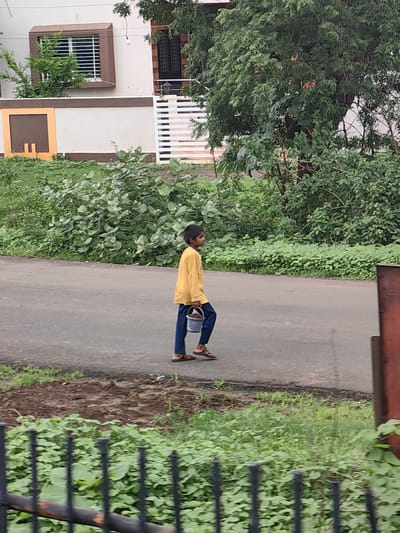 A young boy walks on a paved road in Sakari, India, carrying a light blue bucket in his right hand. He is wearing a yellow long-sleeved shirt and dark blue pants. The road is flanked by lush green vegetation and some trees. In the background, there is a white building with brown trim and a window with a security grate. A black metal fence is visible in the foreground.