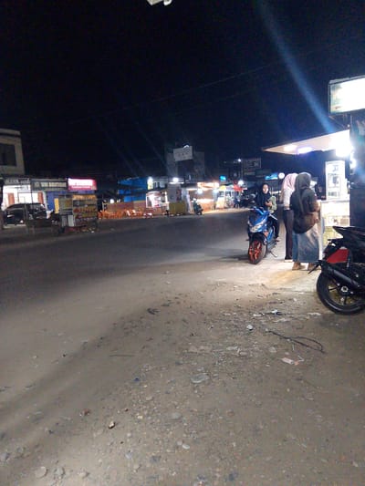 At night in Gampongpaya, Indonesia, a dirt road is illuminated by the lights of various shops and a streetlamp. A blue motorcycle is parked on the side of the road, with a person sitting on it wearing a green jacket and black hijab. Two other individuals, both women wearing hijabs, stand near the motorcycle. One is wearing a pink and white striped shirt and dark pants, and the other is in a dark top and a long grey skirt. A black motorcycle is parked in the foreground, partially obscuring the view. In the background, several food stalls with orange plastic chairs are visible, and a building with the sign "WM BOUTIQUE" is also present. The sky is dark, with a bright beam of light originating from the upper right of the image, possibly from a streetlamp or a sign. The ground is covered in dirt, gravel, and some scattered debris.