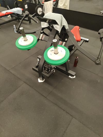The image depicts the interior of a fitness center in Nahariah, Israel. A black plate-loaded exercise machine, equipped with a red padded seat and backrest, is positioned centrally. A light grey towel is draped over the top of the machine's backrest. Two vertical weight plate holders extend from the machine; each holds a larger green weight plate and a smaller white weight plate, the latter displaying "PROF IT" and the number "5." A dark red water bottle rests on the dark grey rubber mat flooring beside the machine's right base. In the background to the left, a second exercise bench with a red pad is visible, draped with a light blue towel, and a clear water bottle is near its base. A large mirror at the rear of the room reflects the lower half of at least two individuals in light-colored attire standing further back, and a sign with the number "8" on a dark surface. The floor throughout the visible area is covered with dark grey interlocking rubber mats. No direct human actions or interactions are depicted in the foreground.