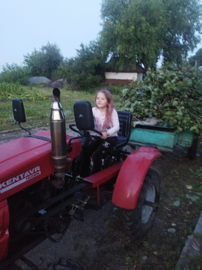 A young girl with pinkish-blonde hair is seated in the driver's seat of a red tractor, appearing to be in Sosnivka, Ukraine. She is wearing a white long-sleeved shirt with a graphic design and dark pants. The tractor has a tall, metallic exhaust pipe on its left side, and two black side mirrors are attached to the frame behind the hood. The word "KENTAVR" and "160BN" are visible in white lettering on the red hood. In the background, there are trees, a building with a white and brown facade, and a pile of green foliage. A teal-colored rectangular object is situated behind the tractor. The ground is a mixture of dirt, grass, and some loose stones. The sky is overcast and light.