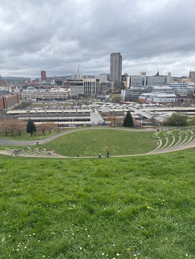 The image shows a view of Sheffield, United Kingdom from a high vantage point. In the foreground is a grassy hillside with walking paths. People are seen walking and standing on the paths and in the grass. The background features a dense urban landscape of Sheffield, including numerous buildings of varying heights and architectural styles. The sky is overcast. The image is oriented sideways.