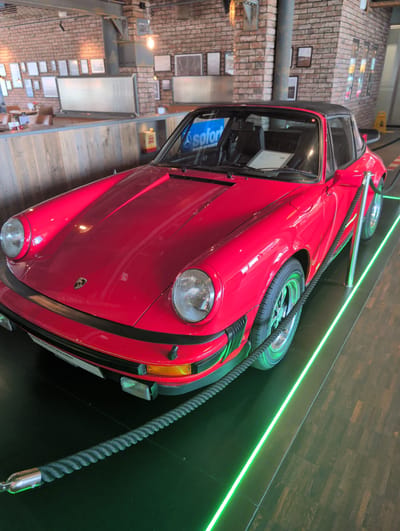 This media file depicts a vibrant red vintage car, possibly a Volkswagen Beetle, displayed in an indoor setting with a brick wall and wooden accents. The car is positioned at an angle, appearing to be suspended or mounted in an unusual display. It features a spare tire mounted on the exterior, a detail often seen on older models. A thick, dark rope with a metallic end is strategically placed alongside the car, perhaps for display purposes or as a safety barrier.

The background includes a brick wall with various framed pictures or signs. There are also shelves and display elements suggesting a museum, exhibition, or a themed retail space. The lighting is a mix of ambient light and prominent green neon strip lights running along a dark wooden wall, creating a modern contrast to the vintage car.

Visible text includes "sofort" on a blue sign inside the car's open door, and partial text on a white sticker on the side of the car, which appears to be German text related to "NACHRICHTEN" or "FAHRZEUG". The scene appears to be well-lit, indicating an indoor environment, likely during daylight hours, though the artificial lighting is dominant. The overall impression is a curated display of a classic automobile, possibly within a location in Böblingen, Germany, as suggested by the context. There are no people visible in the frame.