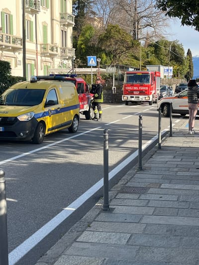 The scene depicts a roadside incident response on a sunny day in Griante, Italy. On the left, a yellow "Anas" utility van with roof-mounted lights is parked on an asphalt road next to a solid white line. Behind it, a red "Vigili del Fuoco" (Fire Department) smaller utility vehicle is partially visible. A firefighter in dark uniform with yellow reflective stripes stands beside this red vehicle, appearing to attend to equipment or an obscured individual. Further down the road, a large red "Vigili del Fuoco" fire truck is parked, facing the viewer. On the far right, a white vehicle marked "Soccorso" (Rescue) is partially visible behind the fire truck. Additional civilian vehicles are queued in the background.
The environment includes a stone-paved sidewalk on the right, lined with dark metal bollards. On the left, across the road, stands a multi-story building with light-colored facades, green shutters, and balconies. Behind the vehicles and buildings, various trees and shrubs are visible, some with bare branches, others evergreen. A blue pedestrian crossing sign is prominent in the midground. Distant hills or mountains are visible under a clear sky in the background. A person in a striped top and light pants stands on the sidewalk on the right, facing away from the camera. Shadows are cast by the vehicles and objects, indicating sunlight from the left.