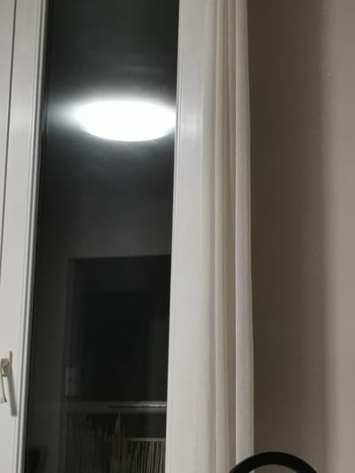 The image shows a dimly lit interior scene, likely a room, viewed through a glass door or window. In the upper portion of the glass, a bright, circular light fixture is visible, reflecting off the glass surface. Below this light, a darker rectangular shape suggests a doorway or a large object. The lower part of the glass reveals a densely packed bookshelf filled with numerous items, appearing to be books or magazines. To the right of the glass area, a white curtain is partially drawn, obscuring some of the view and casting soft folds. The wall to the right of the curtain is a neutral, light brown color. The overall impression is one of an indoor space, possibly a residential or office environment, with a focus on the contrast between the bright light, the dark interior, and the textures of the curtain and the wall. The location in Bourg-lès-Valence, France, is not directly discernible from the visual elements presented in the image itself, as there are no specific geographical markers or recognizable landmarks.