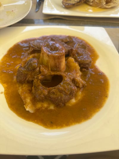 A close-up, overhead shot shows a dish of osso buco, a traditional Italian dish made with cross-cut veal shanks braised with vegetables, white wine, and broth. The veal shanks are cooked until tender and served with a rich, brown gravy. The dish is presented on a large, cream-colored plate, with a portion of what appears to be mashed potatoes or polenta underneath the veal. The lighting suggests it is daytime, possibly outdoors or in a well-lit indoor setting. The setting appears to be a restaurant, with other plates and cutlery visible to the left of the main dish. The focus is entirely on the food, emphasizing its texture and sauce.
