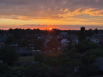An aerial, high-angle, full shot captures a sunset over a rural village in Pratasevichi rural council, Belarus. The sky is a dramatic gradient of orange, yellow, and pink, with wispy clouds catching the last rays of sunlight. Below, the village is silhouetted against the vibrant sky, with the dark shapes of trees and rooftops filling the lower half of the frame. The village appears to be sparsely populated, with houses of varying sizes and styles scattered among the trees. The overall impression is one of peaceful serenity and the natural beauty of the Belarusian countryside at dusk.