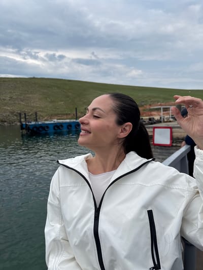 A medium shot shows a woman with dark hair pulled back in a ponytail, wearing a white jacket with a black zipper, and a white t-shirt. She is facing to her left, with her eyes closed and a slight smile on her face, appearing to enjoy the moment. Her right hand is raised slightly near her head. In the background, there is a body of water, a blue boat docked at a pier, and a grassy hillside under a cloudy sky. The scene is likely on Olkhon Island, near the village of Khuzhir, Russia, a popular tourist destination on Lake Baikal.
