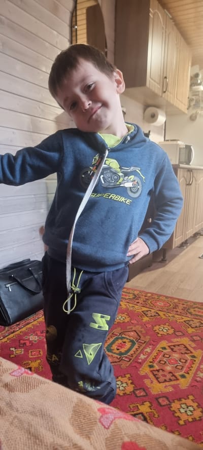 In this media file, a young boy is the sole subject, lying on a bed or sofa. He is wearing a blue hooded sweatshirt with a graphic of a dirt bike and the text "SUPERBIKE" on it, along with dark blue sweatpants featuring yellow triangular designs. The boy is looking directly at the camera with a slight smile, his head tilted.

The setting appears to be indoors, likely a home in Troitskoye, Russia. Behind the boy, a large rug with a traditional red and yellow geometric pattern is visible. To the left, wooden paneling and cabinetry suggest a domestic environment, possibly a kitchen or living area. A roll of paper towels and a black bag are also present in the background. The lighting suggests it could be daytime, though indoor lighting also contributes. The overall mood conveyed by the image is relaxed and informal, with the boy appearing comfortable and at ease.