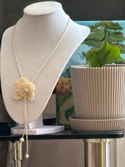 A light-colored necklace with a flower pendant and pearl accents hangs on a white jewelry bust. To the right, a potted plant with green leaves sits on a shelf next to a painting of a tree. The background is dark. There are no people or actions depicted. The scene is indoors and appears to be a still life arrangement. No specific location in Tskhinvali, South Ossetia can be determined from the image.