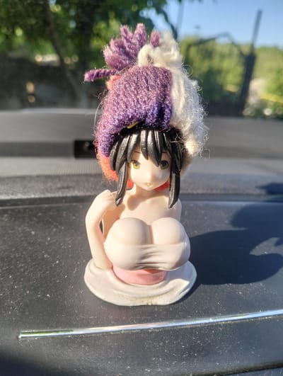 A small figurine of a female anime character with black hair, large breasts, and wearing a knitted hat sits on a dark, textured surface, likely a car dashboard. The figurine has a pale complexion, yellow eyes, and its mouth is slightly open. The hat is multicolored, with sections of purple and white yarn, and appears to be adorned with fluffy tufts. The background is blurred, showing greenery and a bright sky, suggesting an outdoor setting, possibly in or near the city of Șoldănești, Moldova. A thin, reflective strip runs horizontally across the foreground. The lighting indicates it is daytime, with shadows cast by the figurine.