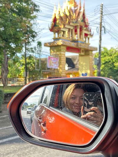 A woman is taking a selfie in the side-view mirror of an orange car. The mirror reflects her face, her phone, and her hand holding the phone. She is smiling and looking at the camera. The background of the reflection shows a street scene with other cars and trees. Behind the car, an ornate golden structure with a red roof is visible. This structure appears to be a gate or a shrine, characteristic of religious or cultural landmarks in Ao Luek, Thailand. The sky is partly cloudy. The overall scene suggests a travel or road trip moment in a tropical setting.