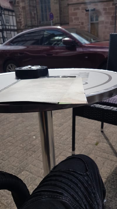 A ground-level, rotated outdoor shot shows a black chair partially visible to the left and another chair to the top. A table with a white top and black rim is on the left, reflecting the reflection of a maroon car and stone building. A sign with a red and blue circle stands in the reflection of the table. To the right is a person wearing black jeans and sitting in a chair. The ground appears to be concrete. This location may be in the city of Rotenburg an der Fulda, Germany.
