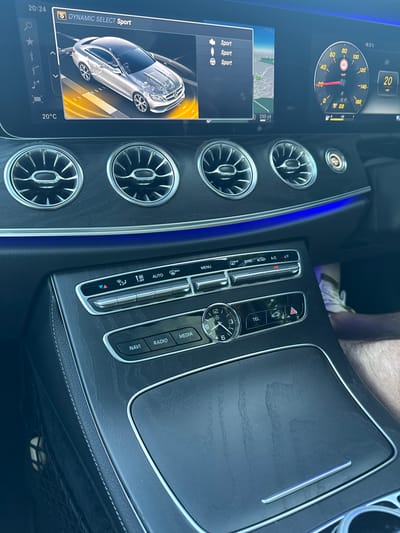 The image shows the interior of a car, likely a Mercedes-Benz, with a focus on the dashboard and center console. The scene is captured from a slightly elevated perspective, looking down and to the right.
The dashboard features a digital instrument cluster displaying a speed of 20 mph and the outside temperature at 18.5°C. Below the cluster, a navigation screen is visible, showing a map with a distance of 200 yards. The screen also displays "S DYNAMIC SELECT Sport" and the time "20:24". Several circular air vents are prominent on the console.
The center console includes a series of buttons and a digital display. Visible controls are labeled "MENU," "AUTO," "A/C," "SYNC," "TEL," "NAVI," "RADIO," and "MEDIA." A classic analog clock is integrated into the console, showing the time as approximately 4:05. The interior is illuminated by ambient blue lighting, suggesting it might be evening or that the interior lighting is activated.
The background shows a portion of a person's leg and arm, indicating a driver or passenger is present in the vehicle. The setting appears to be a car parked in or driving on a road, given the navigation display and speedometer. No specific weather conditions are evident, but the temperature suggests it is not extremely cold. The overall impression is a modern, well-equipped car interior.