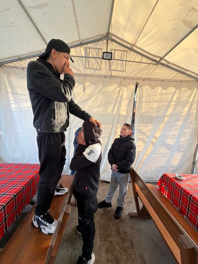 Three young males are sitting on benches inside a tent. The bench is covered with a red-and-black checkered tablecloth. The tent has a white fabric cover with a metal frame. The location is Zlatna Panega, Bulgaria. One male is wearing a black hat, a black jacket, and black pants. He is holding his hand near his mouth. Another male is wearing a black hooded sweatshirt with a white patch on the sleeve, black pants, and black and white sneakers. He has his hand on his head. The third male is wearing a black puffy jacket and grey pants.