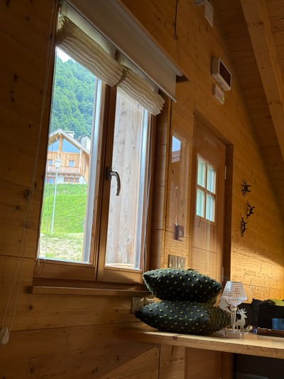 The image depicts the interior of a room with wood-paneled walls and ceiling, likely in a mountain chalet in Sauris/Zahre, Italy. A window on the left reveals a verdant hillside with a wooden house in the distance. On a wooden shelf in the foreground, two dark green decorative pillows with light green polka dots are stacked. To the right of the pillows, a crystal table lamp with a diamond-like pattern sits on a base. A small white reindeer figurine is also visible next to the lamp. The wall to the right of the window has three black metal wall hooks and a small white sign with green text. A digital display with red text is mounted higher on the wall, above a white rectangular object.