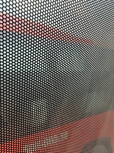 The image is a close-up, angled view of a perforated surface, likely a window or screen. The perforations are circular and arranged in a regular grid pattern. Through the holes, a background is visible, featuring a diagonal band of bright red or orange color against a lighter, possibly white or pale yellow, area. The red/orange area shows some indistinct, blurry shapes that could be text or graphics. The lighting suggests this is an outdoor scene, possibly a window of a vehicle or building in Dĩ An, Vietnam, where such materials are commonly used for advertising or privacy screens on shops and public transport. No people or specific identifiable objects are clearly visible due to the focus on the perforated pattern and the blurry background.
