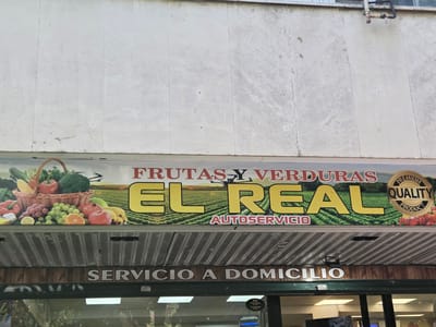 This is a medium shot, eye-level, exterior photograph of a fruit and vegetable stand located in Collado Villalba, Spain. The sign for the stand, which reads "FRUTAS Y VERDURAS EL REAL AUTOSERVICIO", is prominently displayed across the top of the frame. The sign features large yellow lettering for "EL REAL", with "FRUTAS Y VERDURAS" above and "AUTOSERVICIO" below. To the right of the main text is a circular emblem with the words "PREMIUM QUALITY PRODUCT" and an image of red peppers. The background of the sign depicts a green field with a drone flying overhead, and a basket filled with various fruits and vegetables such as broccoli, tomatoes, grapes, bananas, oranges, and lemons. Below the main sign, another banner reads "SERVICIO A DOMICILIO" which translates to "Home Delivery". The stand itself appears to be an open-fronted structure with a wooden interior, and a green counter is visible at the bottom of the frame. Natural lighting illuminates the scene, casting minimal shadows. There are no people visible in the image.