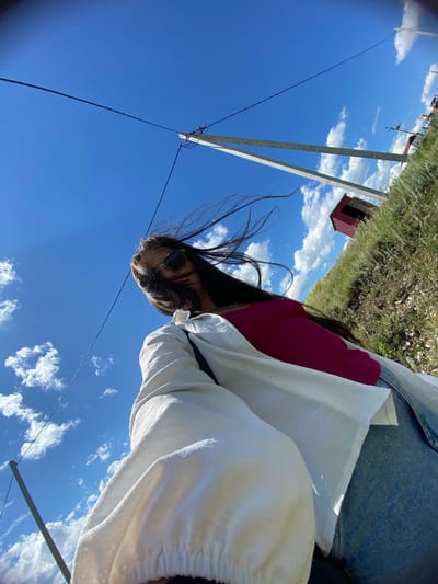 A low-angle, wide-angle shot captures a young woman standing outdoors in Yuldybayevo, Russia, against a bright blue sky with scattered white clouds. The woman, with long dark hair blowing in the wind, wears sunglasses, a pink top, and a white oversized shirt. She is holding a phone, presumably taking a selfie. In the background, a grassy hillside is visible, dotted with small shrubs and a small red building. Power lines stretch across the frame, supported by utility poles, indicating a rural or semi-rural setting. The overall impression is one of a casual, sunny day.