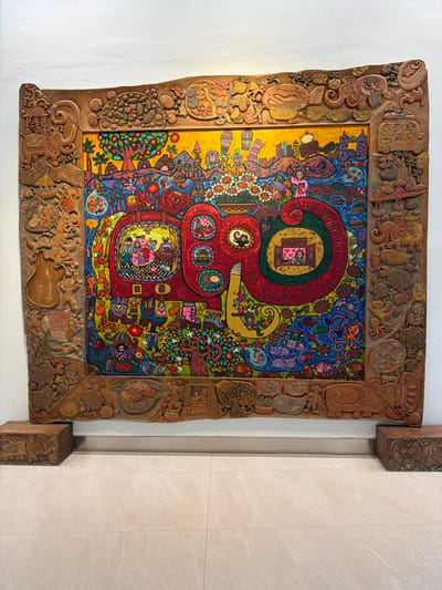 This is a close-up, high-angle shot of a vibrant, intricate artwork housed in a carved wooden frame. The artwork itself is a colorful and surreal painting with a dreamlike quality, featuring stylized figures, fantastical creatures, and a whimsical cityscape. The painting is dominated by a large, red, spiral-like shape, which contains a scene of people in a bed, reminiscent of a storybook illustration. Surrounding this central motif are numerous small figures, animals (including octopuses and fish), and swirling patterns in blues, greens, and yellows. The detailed wooden frame surrounding the painting is also heavily carved with various symbolic motifs, including human faces, animals, and abstract shapes, suggesting a cultural or artistic significance. The overall impression is one of imaginative storytelling and meticulous craftsmanship. The artwork is mounted on a white wall and is adjacent to a beige tiled wall, with two wooden pillars flanking the artwork, one on each side. The lighting appears to be interior, casting a soft, even illumination on the scene. There are no discernible time-of-day or weather cues. The location context provided is Rawa Burung, Indonesia, which might inform the interpretation of the symbols within the artwork and its frame, though specific identifiable features within the image do not directly confirm this location.
