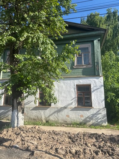 A two-story wooden house, painted dark green on the upper floor and white on the lower, stands partially obscured by a large tree with lush green foliage. The house has multiple windows with wooden frames. The lower portion of the house has a rough, textured white finish, with some visible damage. The upper floor is clad in horizontal green wooden planks, with a projecting roofline. A pile of brown soil is in the foreground, obscuring part of the sidewalk and a portion of the lower wall of the house. Power lines are visible against a clear blue sky, with colorful bulbs strung between them. The scene appears to be in an urban or suburban setting in Vereya, Russia, with a hint of rural charm due to the natural elements.