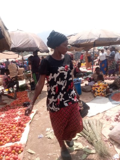 This image captures a vibrant and bustling open-air market scene, likely during daylight hours, characterized by bright sunlight and numerous people.
In the foreground, a woman with dark skin and a dark headwrap walks towards the right side of the frame, her head turned slightly to her right, looking into the distance. She wears a black short-sleeved top with a prominent white, red, and black abstract or geometric pattern on the front. Her lower garment is a patterned red and black wrap skirt or trousers. She carries a dark-colored bag or item in her left hand, which is partially visible. Her right arm is extended slightly forward. She appears to be wearing dark, open-toed shoes.
To the left of the woman, a significant quantity of fresh red tomatoes is laid out on a light-colored ground covering, possibly a tarp or mat, indicating a produce stall. Some darker red, smaller fruits or berries are also mixed in with the tomatoes. The ground throughout the market appears to be dirt or earth.
The background is densely populated with market activity. Several large, light-colored (beige or white) umbrellas provide shade over various stalls. Under these umbrellas and in other areas, numerous individuals are visible – vendors attending to their goods and customers browsing. Some people are seated on the ground among their produce. For example, to the right of the main woman, another person is seated near a spread of what appear to be red onions or similar vegetables. Further back, more market-goers and vendors, many dressed in colorful patterned clothing, contribute to the busy atmosphere.
Near the bottom right corner of the image, on the ground, there is a bundle of dried plant material, possibly brooms or traditional crafting materials. The overall impression is that of a lively community market where various goods, primarily fresh produce, are bought and sold. No discernible text is visible in the image.