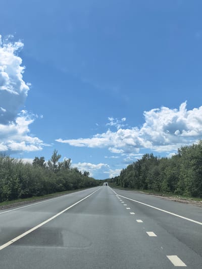 The image depicts a highway stretching into the distance under a bright blue sky with scattered white clouds. The road has two lanes in each direction, separated by a solid white line. The lanes are marked with dashed white lines. On either side of the road, dense green trees form a continuous line. In the distance, a white truck is visible on the highway. The scene appears to be a rural or suburban area, possibly on the outskirts of Vozrozhdenie, Russia, given the natural landscape and the highway infrastructure. There are no people or specific actions captured in the frame.