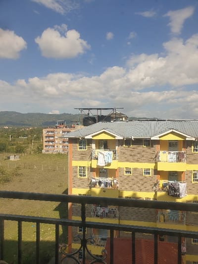 The image displays an exterior daytime scene in Mumbuni North ward, Kenya. In the immediate foreground, a dark metal balcony railing, featuring decorative scrollwork, partially obscures the lower portion of the view. The primary subject in the midground is a multi-story residential apartment building with a distinctive facade. This building features patterned brown-grey exterior wall sections interspersed with bright yellow horizontal bands and window frames. Numerous balconies are visible across its elevation, many equipped with clotheslines from which various items of laundry, including shirts, pants, and towels of assorted colors, are hanging to dry. The building is topped with a grey corrugated metal gabled roof, and a lower orange-tiled roof section is also partially visible.
To the left of this building, an expansive, undeveloped plot of land is visible, covered in dry grass. A rudimentary wire fence line runs across this field, and a small, square concrete structure, possibly a utility box or wellhead, is situated within it. In the further background, another multi-story residential building, appearing to be an apartment complex, stands against a backdrop of distant, verdant hills. Above the roofline of one of the more distant buildings, a metal framework supporting two large, dark-colored water tanks is prominently visible. The sky overhead is clear blue with scattered white cumulus clouds. No individuals are visible within the scene.